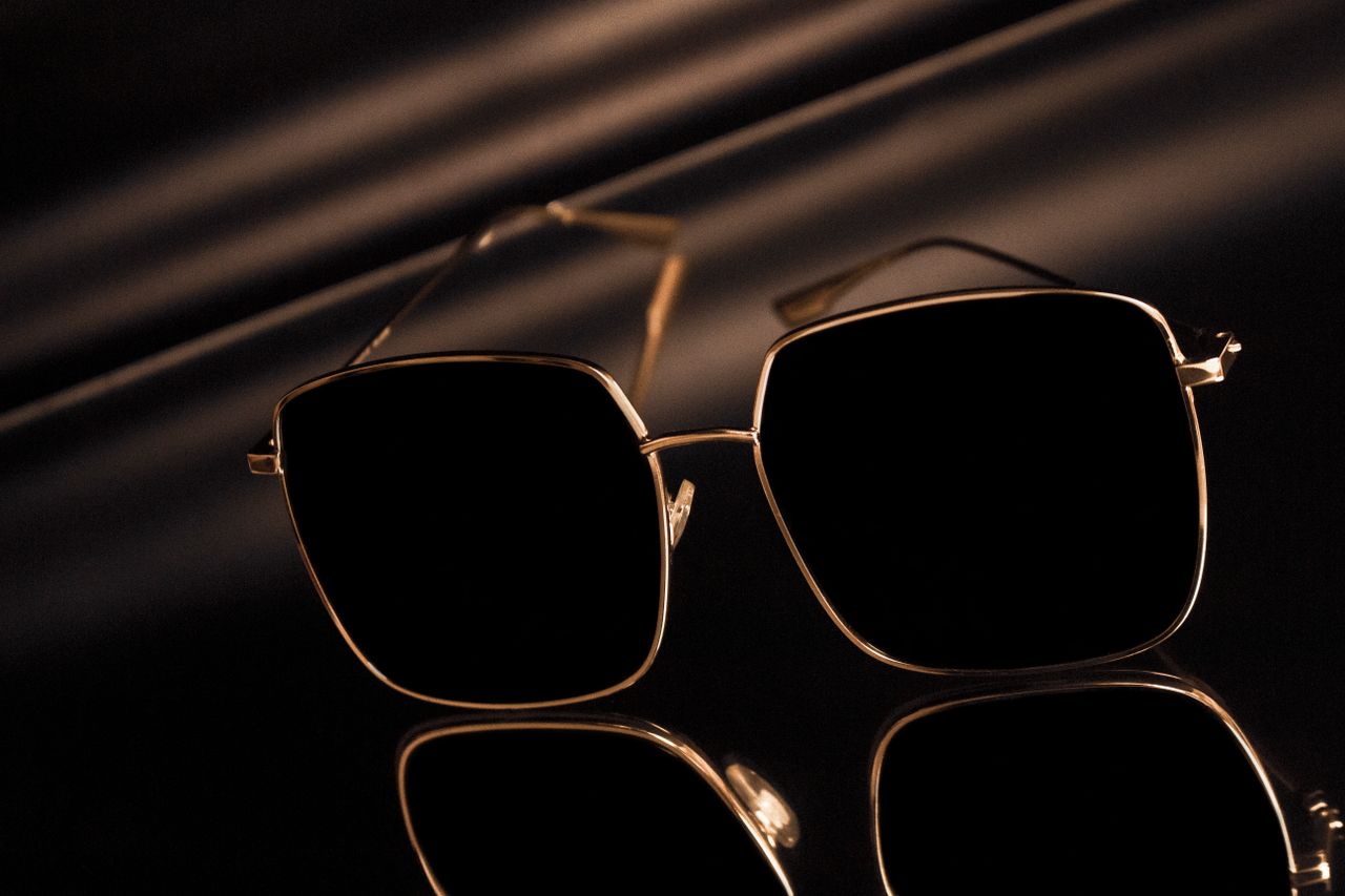 services-04 Stylish square sunglasses with gold frames reflecting on a glossy surface.